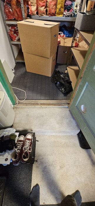 Förråd med kartonger på hyllor, betongtrapppsteg och dörröppning mot rum med skor och matta.