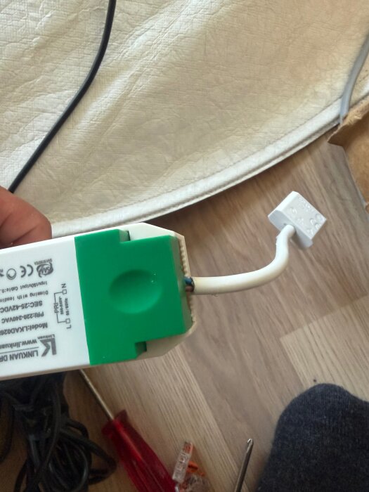 Elektrisk koppling med grönt lock och vit kabel, omgivna av verktyg på ett trägolv.