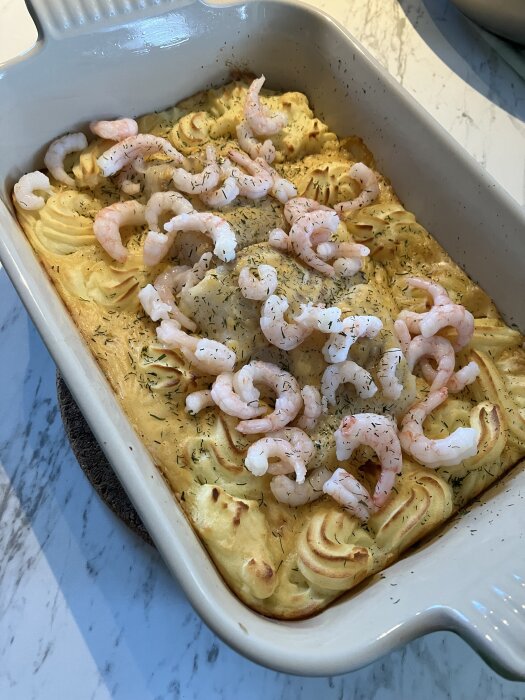 Fiskgratäng i ugnsform med spritsat potatismos, toppad med räkor och dill.