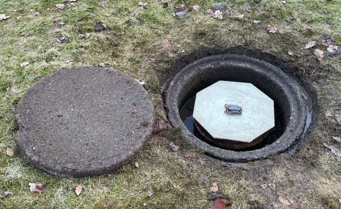 Betonglock och avloppsbrunn på en gräsmatta med ett borttaget betonglock som visar en tank med ett inre täcklock.