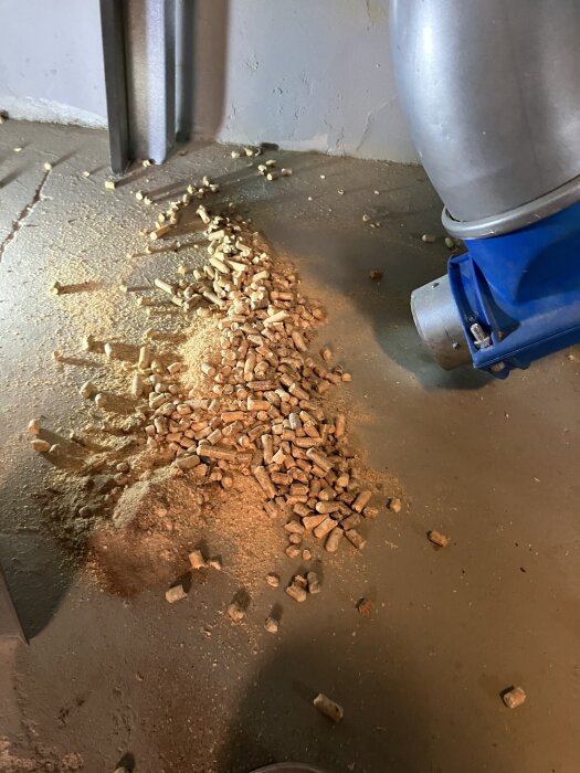 Spilld träpellets och sågspån på ett golv, bredvid en industriell blå och silverfärgad maskin.