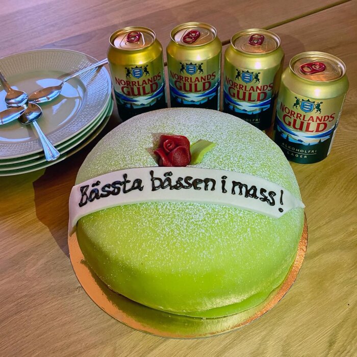 Prinsesstårta med text "Bästa bassen i mas!", dekorationsros och fyra burkar Norrlands Guld, bredvid stapel med tallrikar och skedar.