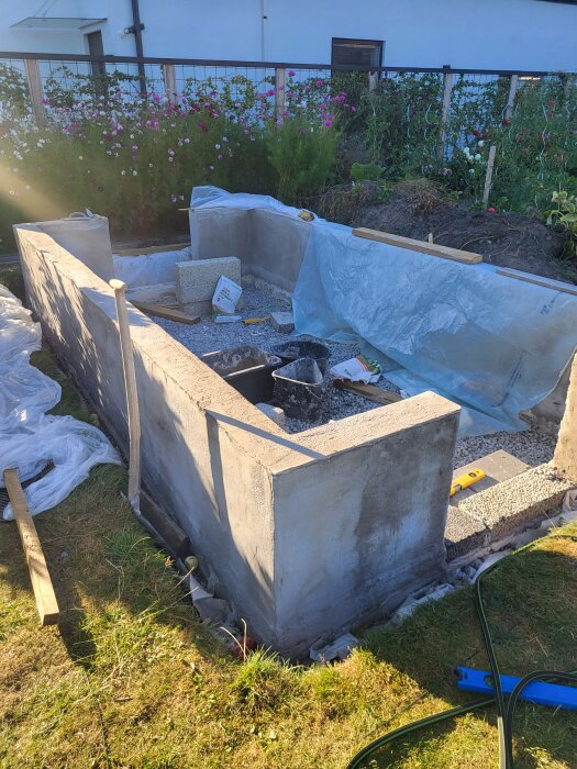 Växthusunderbyggnad med betongväggar, makadam och verktyg i trädgårdsmiljö, omgiven av blommor och byggmaterial.