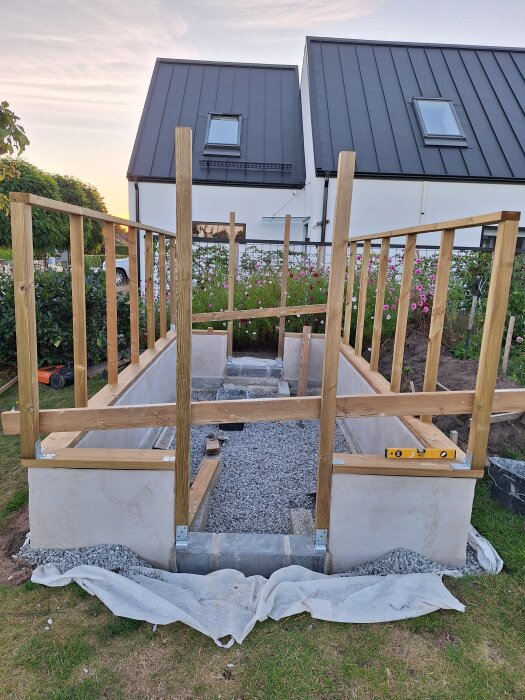 Träramkonstruktion på grusad yta vid sidan om ett hus i trädgårdsmiljö; byggprojekt påbörjat.