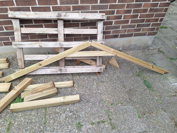 Träkonstruktion med takstol och träbitar på stenlagd uppfart, intill en tegelvägg.