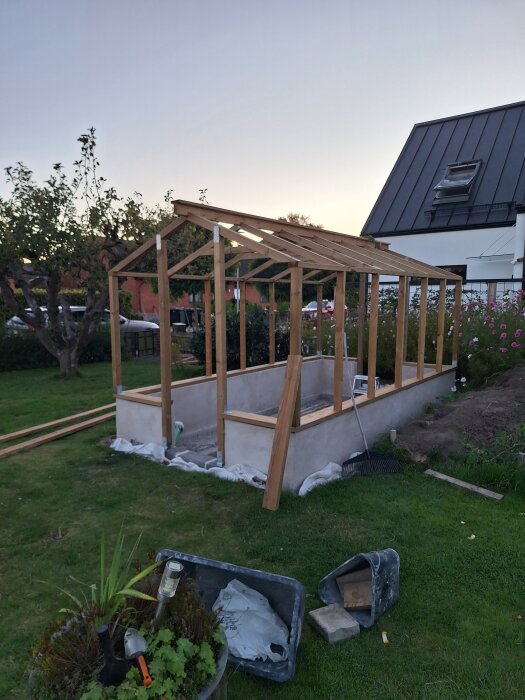 Träram för ett litet byggprojekt på en gräsmatta, omgiven av träd och blommor. Delar och verktyg syns i förgrunden.