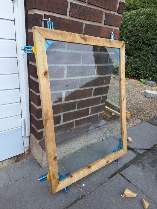 Fönsterram av tunt trä med glas, lutad mot tegelvägg och fäst med blå och gula klämmor, monterad på en stenbelagd uteplats.