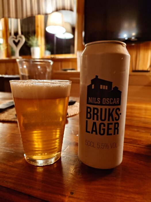 Ett glas med skummande öl bredvid en burk märkt "Nils Oscar Brukslager" på ett träbord i ett mysigt rum.