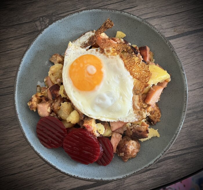 Tallrik med pytt i panna av korv, köttbullar, köttfärsbiffar, potatis, lök, stekt ägg och rödbetor, på ett träbord.