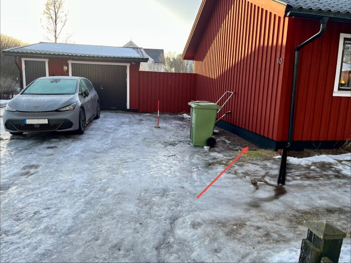 Röd husvägg vid isigt garageuppfart, gröna soptunnor, och pilar visar vattenansamling nära väggen.