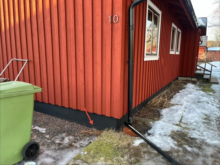 Röd husvägg med vattenansamling vid soptunnor på grusmark, pil visar nivå. Snö och is på marken.