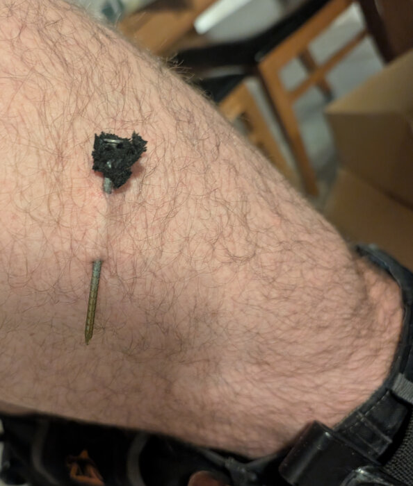 En spik syns instucken i en hårig arm, vilket illustrerar en mindre olycka vid ett byggprojekt.