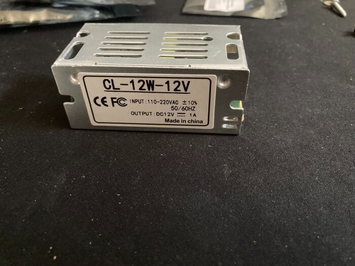 Kompakt nätaggregat CL-12W-12V, in 110-220V AC, ut 12V DC, 1A, CE-märkt, liggande på svart yta. Made in China.