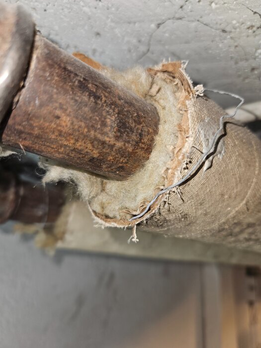 Gammalt värmerör i källartak med delvis borttagen isolering som glittrar vid belysning, visar rör och skadad isolering närbild.