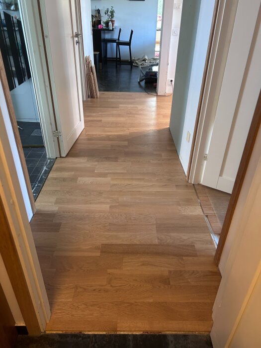 Korridor med nyligen installerat trägolv, anslutning till rum med mörkare golv, dörrar står öppna och byggmaterial syns i bakgrunden.