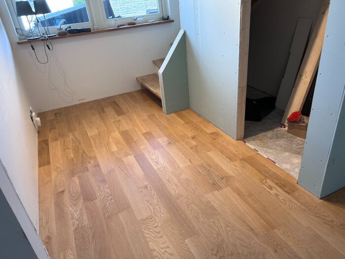 Nylagt trägolv i ett rum under renovering, med en liten trappa och öppning till ett annat rum. Fönster och diverse verktyg syns på fönsterbrädan.
