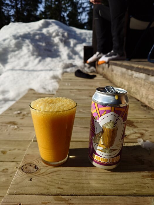 Glas med skummande guldryck och en burk märkt "Smoothie" placerade på ett träbord utomhus, snö i bakgrunden.