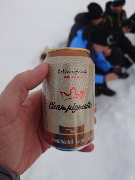 En hand håller en ölburk med texten "Champigneulles Continental Lager" på snöig mark med personer i bakgrunden.