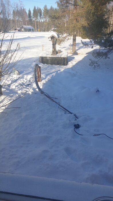 Snötäckta mark och trädgård med en brunn och en nedfallen stolpe med kablar. Träd i bakgrunden och ett hus syns på avstånd.