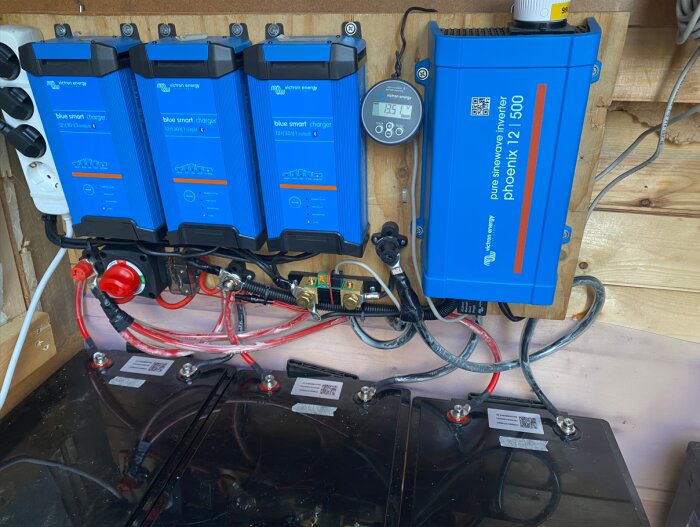 Tre blå batteriladdare monterade på väggen med kopplade kablar och display, bredvid en blå inverter i ett trångt utrymme.