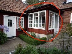 Rött hus med en vit dörr och fönster. En utbyggnad med många fönster är markerad med röd cirkel. Välskött trädgård med stenlagd gång och grönska.