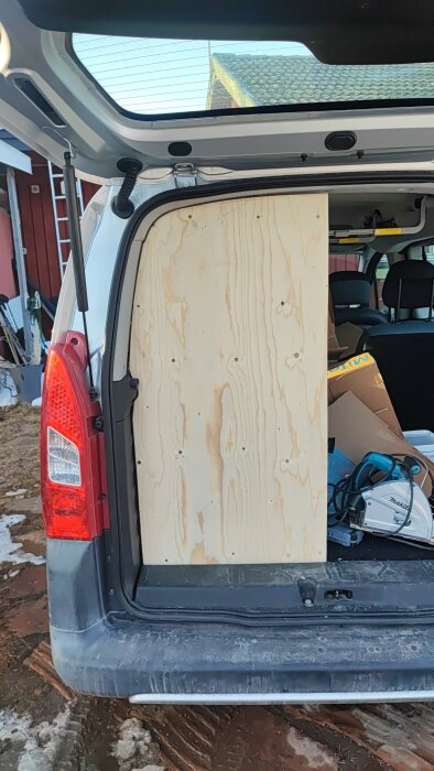 Baklucka på bil med plywoodskiva monterad i öppningen, kringliggande verktyg syns inuti tillsammans med pappkartonger.