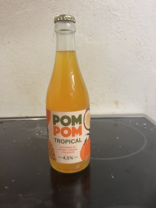 Flaska med etikett "POM POM TROPICAL", smaken är mango, ananas och kokos, alkoholhalt 4,5%, står på en spis.