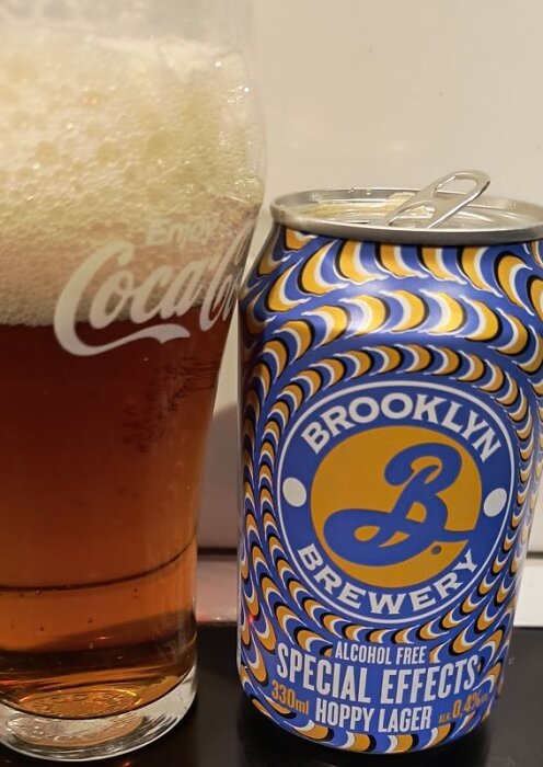 Alkoholfri ölburk från Brooklyn Brewery bredvid ett fullt glas med skum på toppen, delvis med loggan "Enjoy Coca Cola" synlig.