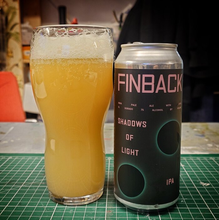 Glas med ljus öl och en burk märkt "Finback Shadows of Light IPA" på ett rutigt underlag.