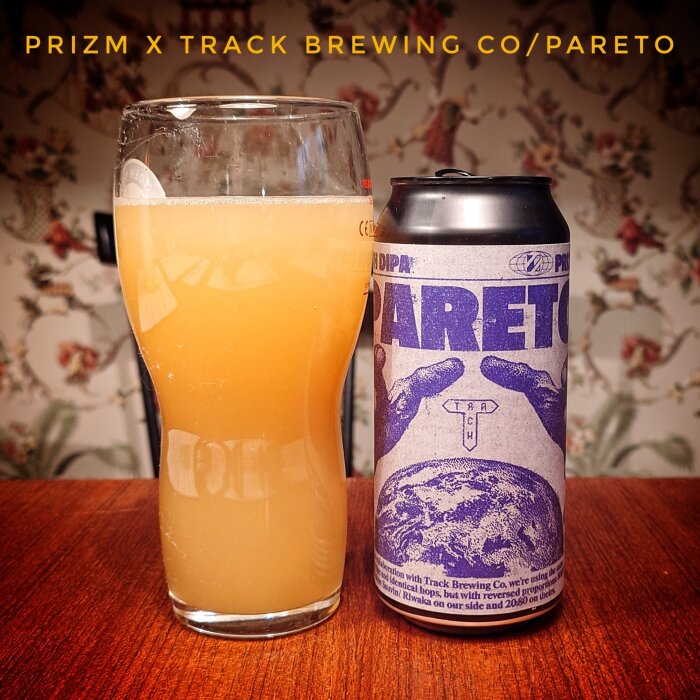 Ett glas med öl bredvid en burk märkt med "Prizm x Track Brewing Co/Pareto" på ett bord med blommigt tapet i bakgrunden.
