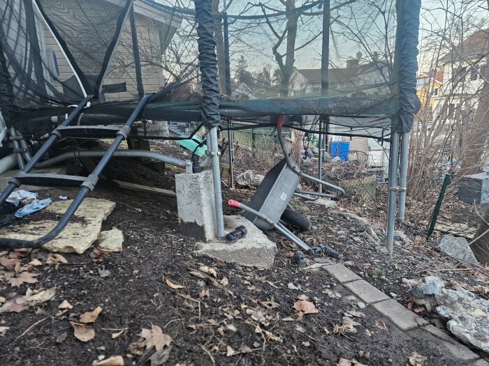 Trädgård med en studsmatta och en skottkärra under den, omgiven av bråte och stenar. Står nära ett hus med träd i bakgrunden.