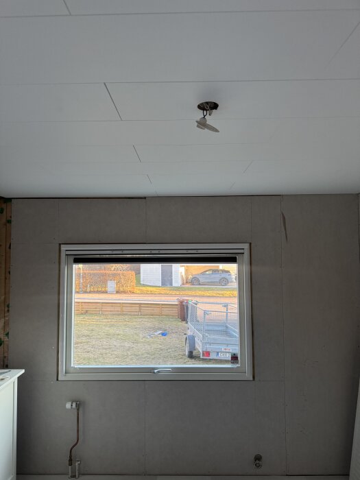 Rum med gipsade väggar, ett stort fönster med utsikt över trädgården och en oinstallerad taklampa; påbörjad elomdragning syns.