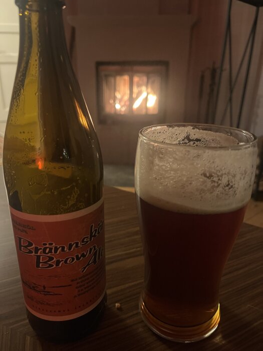 Flaska och glas Brännskär Brown Ale på ett bord framför en tänd brasa.