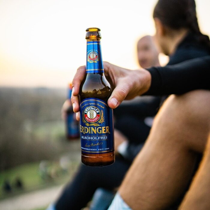 En person håller en flaska alkoholfri Erdinger-öl utomhus med suddiga människor i bakgrunden.