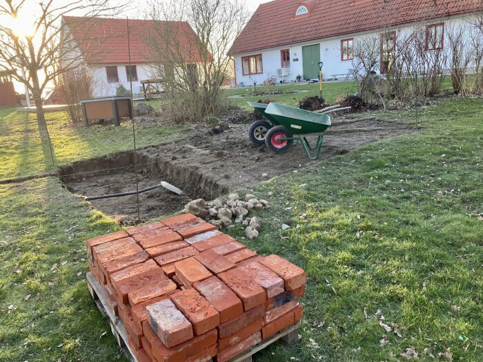 Skottkärra och staplade tegelstenar vid grundarbete för byggprojekt på trädgårdstomt med byggnader i bakgrunden.