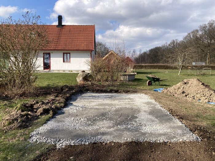Bygget av en grund framför ett vitt hus med rött tak och en skottkärra på gräsmattan, omgiven av träd och buskar.