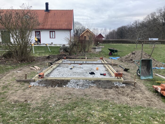 Grus och träbalkar på en inhägnad yta förbereder grunden till ett växthus, med hus och trädgård i bakgrunden. Hjulbörr, tegelstenar och verktyg runtom.