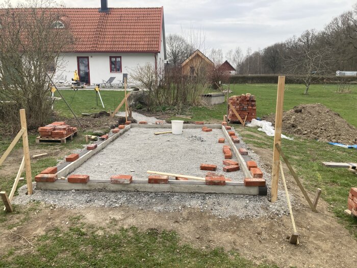 Grundläggning av växthus med ramar och staplade tegelstenar på en grusyta, mot en bakgrund av gräsmatta och hus.