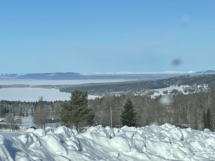 Snötäckt landskap med skog, små hus och en sjö i bakgrunden under klarblå himmel.