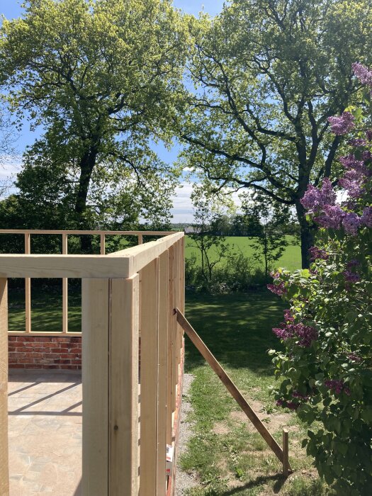 Träram på tegelgrund under uppbyggnad, omgiven av gröna träd och lila blommor, med skog och gräsmatta i bakgrunden en solig dag.