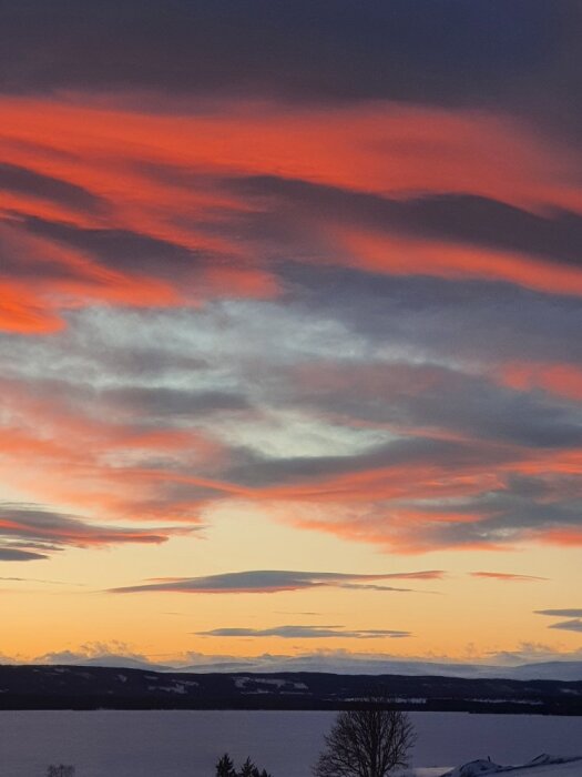 Solnedgång med röd-orange moln mot en mörk landskapshorisont och en sjö i förgrunden.
