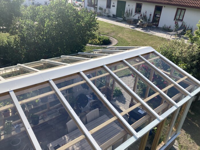 Växthus med glaspaneler och trästomme i en trädgård, omgiven av grönska och utemöbler framför ett hus.
