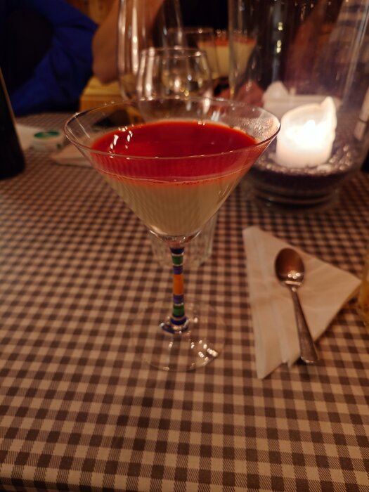 Pannacotta med hallonspegel serverad i ett cocktailglas på en rutig duk vid en dukad bord med sked och servett.