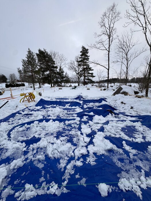 Byggarbetsplats täckt med blå presenningar och snötäckt mark, omgiven av träd.