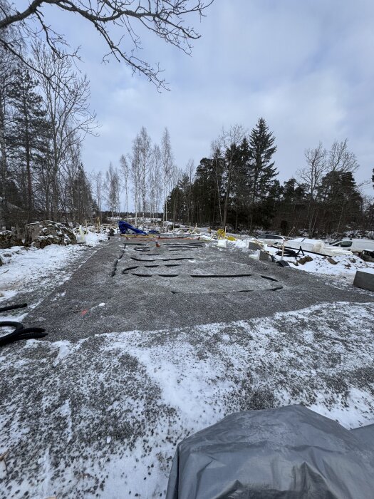 Byggarbetsplats på snötäckt mark med grus och presenningar, omgiven av träd. Grunden är förberedd för ytterligare arbete.