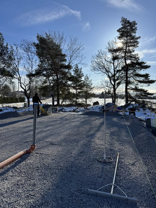 Grusyta med snöfläckar och en person som använder en paddningsmaskin, omgiven av träd och klarblå himmel i bakgrunden.
