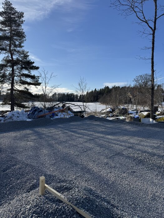 En byggarbetsplats med grusunderlag, omgiven av snötäckta marker och träd under en klarblå himmel.