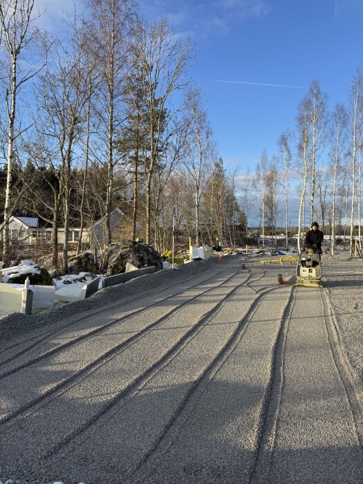 Byggarbetsplats med grusad mark, en man med vibrationstäft står i bakgrunden. Omgiven av barträd och klarblå himmel.