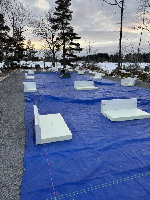 Vy över en bygggrund med blå presenning och vita kantelement av XPS placerade ovanpå, omgiven av snö och träd i vinterlandskap.