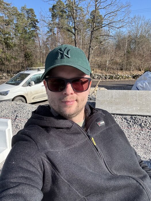 En person i solglasögon och grön keps tar en selfie utomhus, med bil och träd i bakgrunden, en solig vårdag.